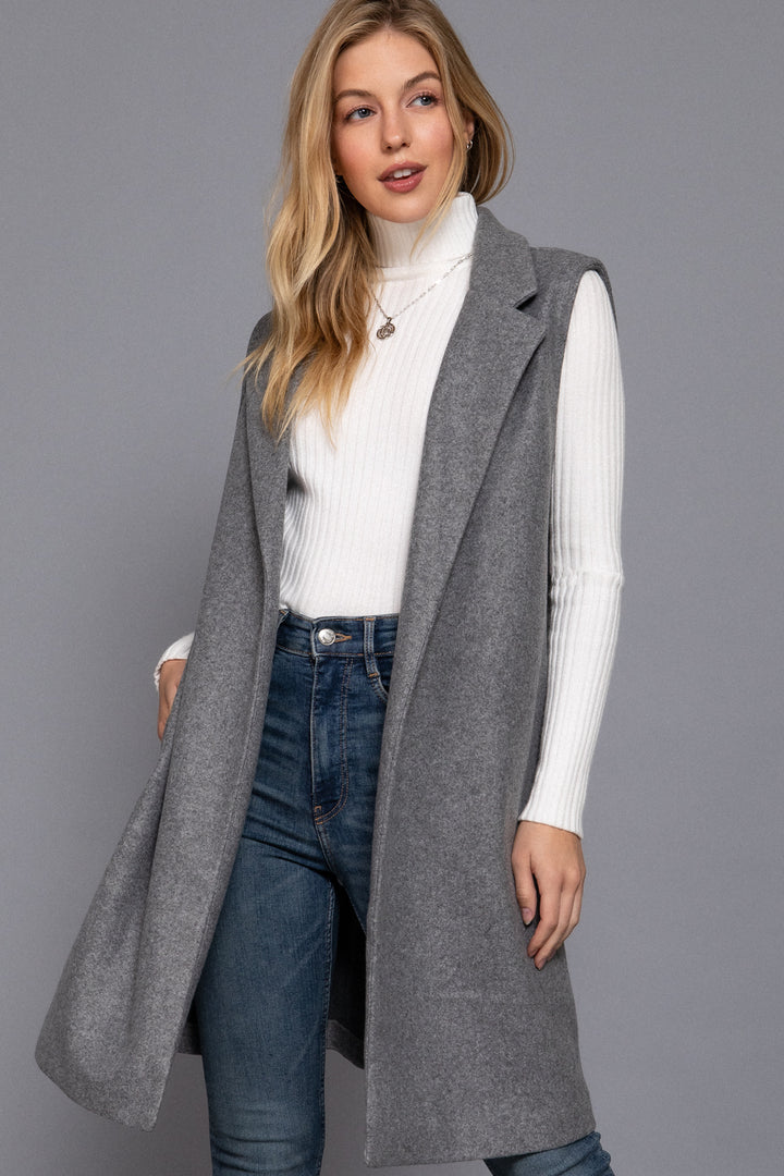 Woman wearing a long gray coat over a white sweater and blue jeans on a gray background