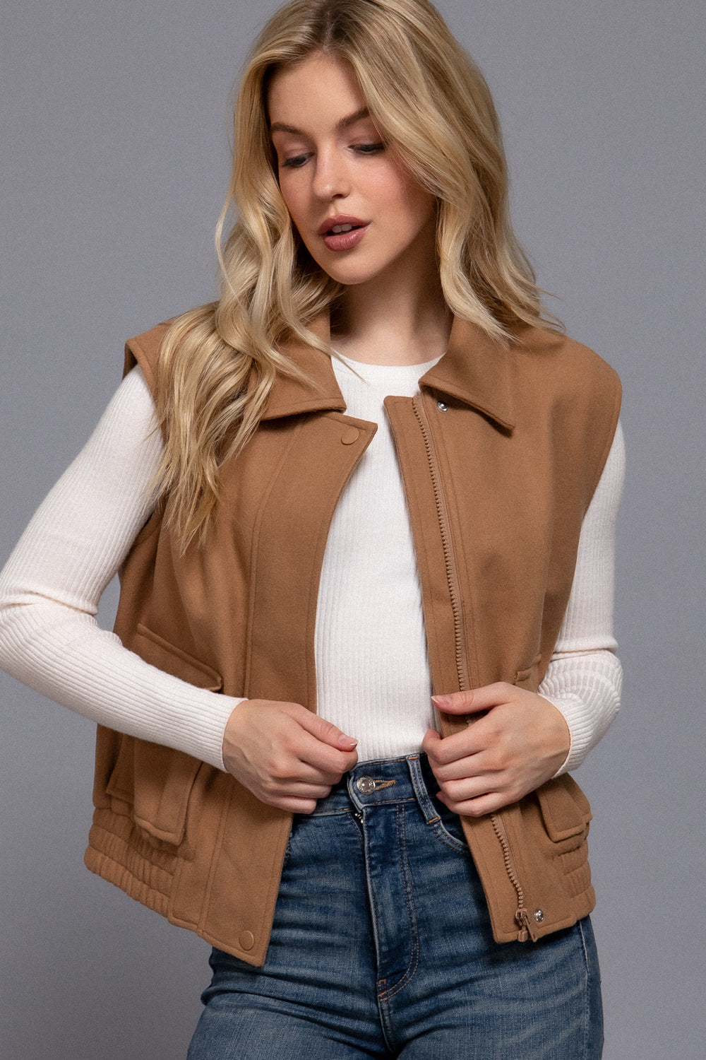 Woman wearing a brown vest over a white shirt and blue jeans on a gray background